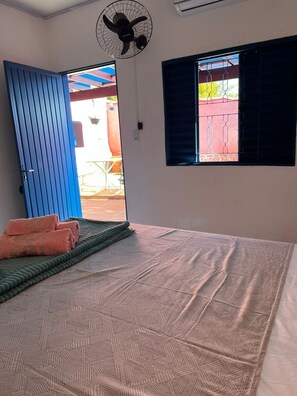 Une chambre avec un lit, un ventilateur, une fenêtre avec des stores et une vue sur l’extérieur.