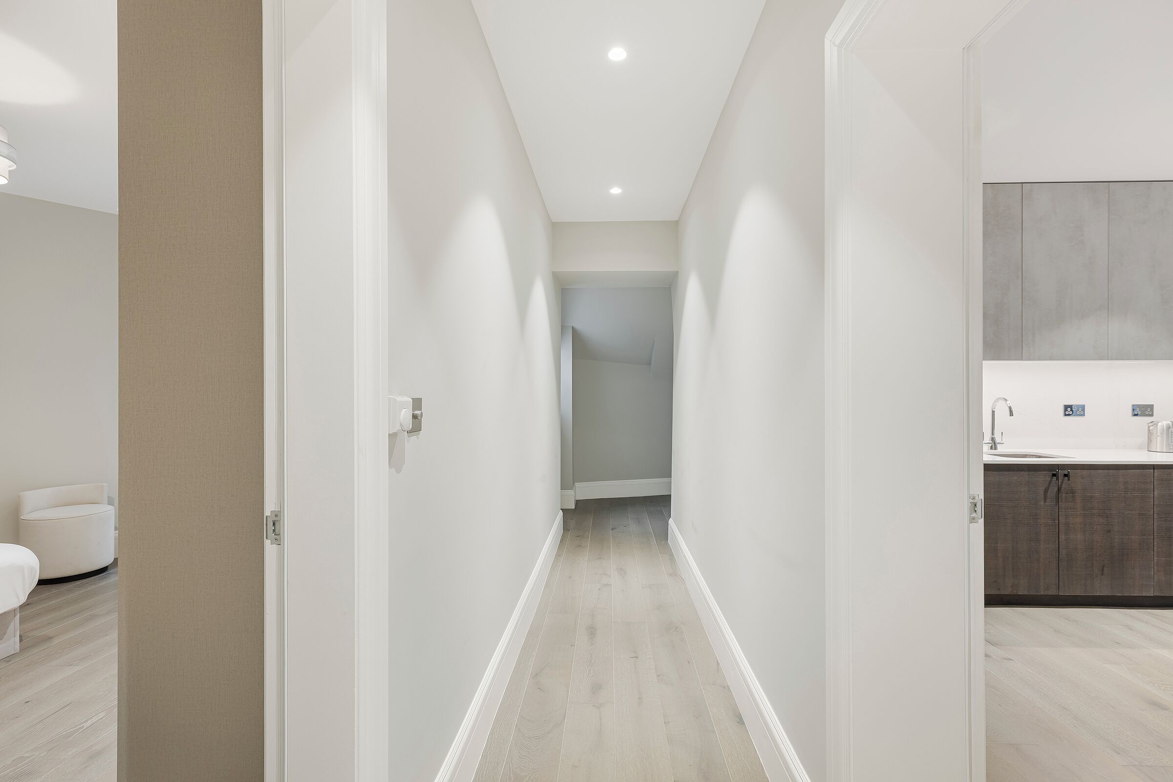 A modern hotel corridor with a neutral color scheme, a wooden floor, and a bathroom with a large mirror and dark cabinets.
