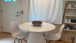 Un comedor con una mesa redonda, cuatro sillas blancas, un cuenco de frutos secos, una lámpara colgante, una cortina blanca y estantes con utensilios de cocina.