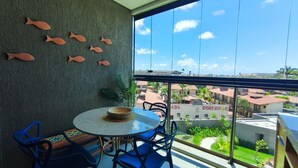 Un balcón con una mesa redonda, sillas azules, un muro decorativo con formas de peces y vistas a edificios y follaje.