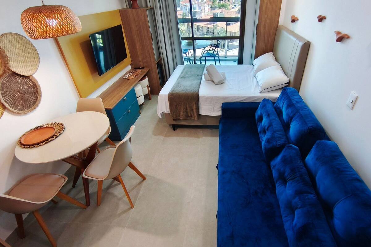 Une chambre d’hôtel moderne avec un lit, un canapé bleu, un coin repas avec une table et des chaises, une télévision et une vue sur l’extérieur.