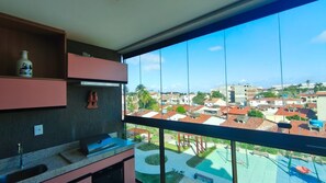 Un balcon donnant sur un quartier résidentiel, une cuisine équipée d’un évier et d’un barbecue, et une étagère sur laquelle se trouvent une bouteille et une paire de chaussures.