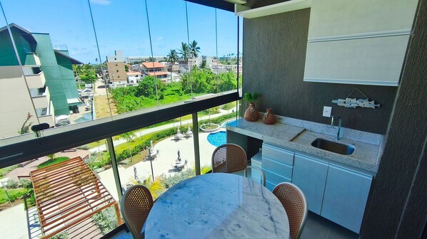 Un balcón con vista a una piscina, un comedor con mesa y sillas, y una cocina con pileta y armarios.