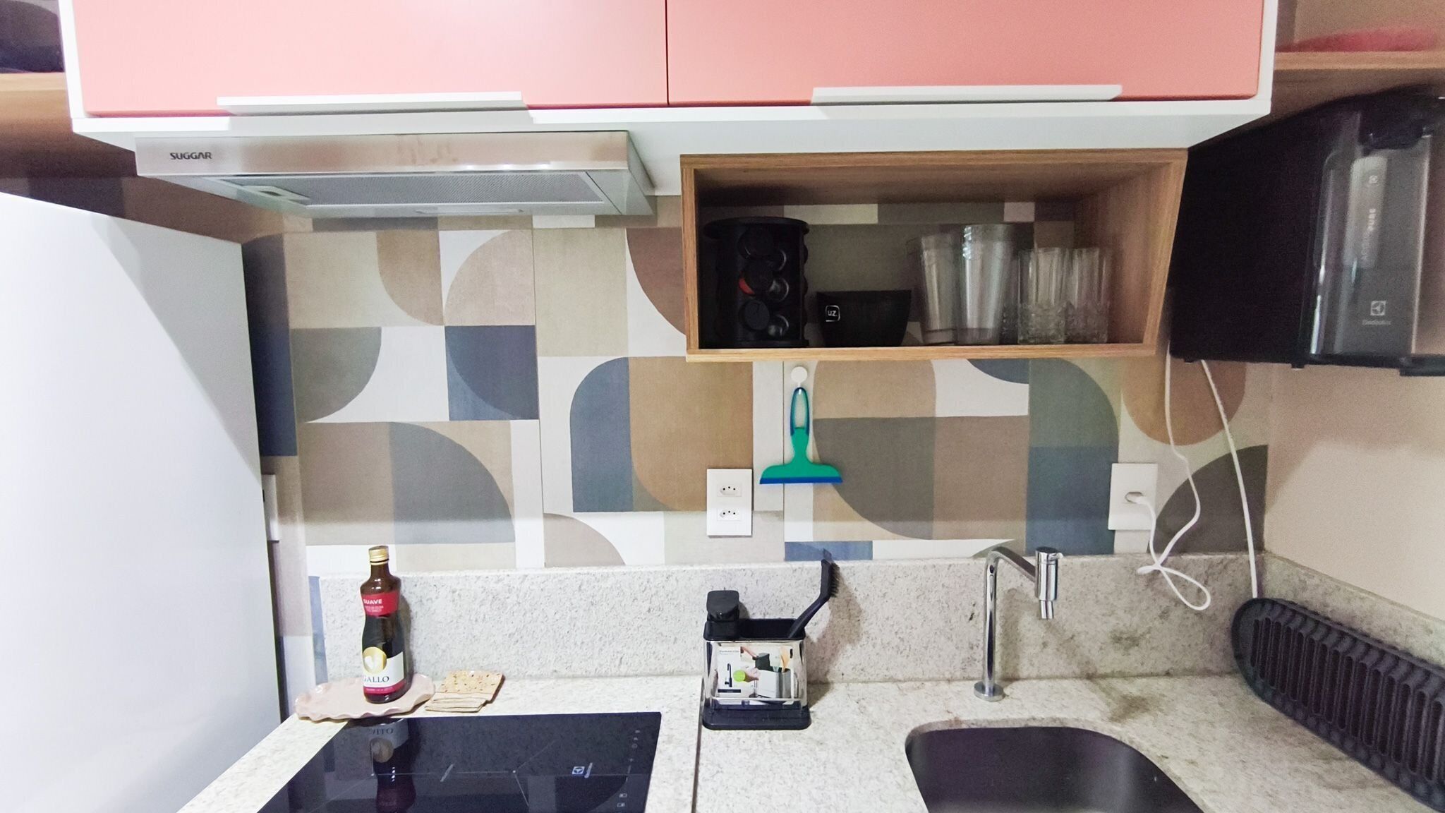 A compact kitchen with a microwave, a small shelf with kitchenware, a bottle of wine, and a sink.