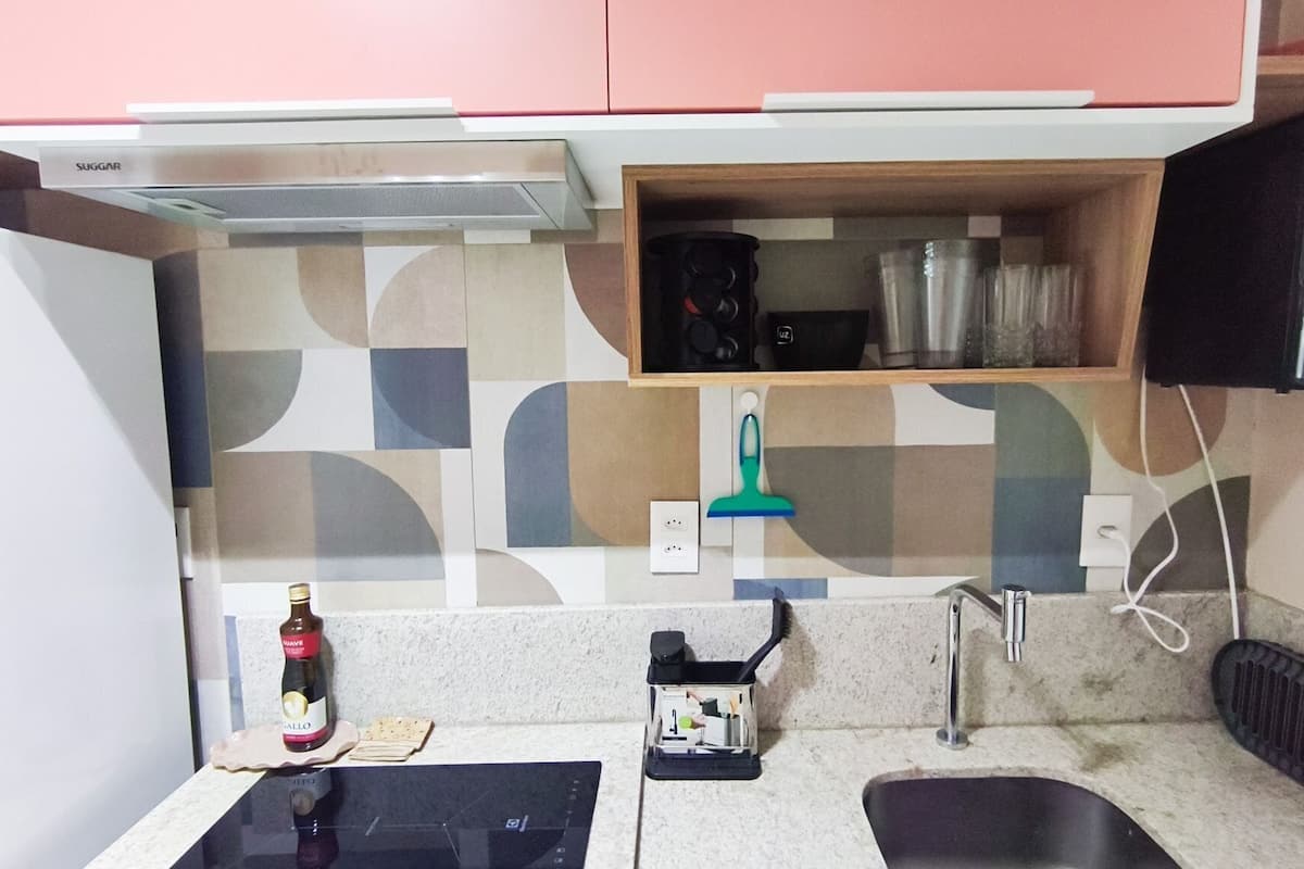 A compact kitchen with a microwave, a small shelf with kitchenware, a bottle of wine, and a sink.