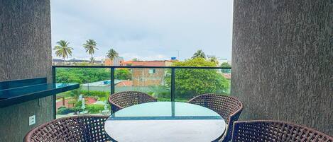 A balcony with wicker furniture and a glass table, overlooking a cityscape with buildings and palm trees.