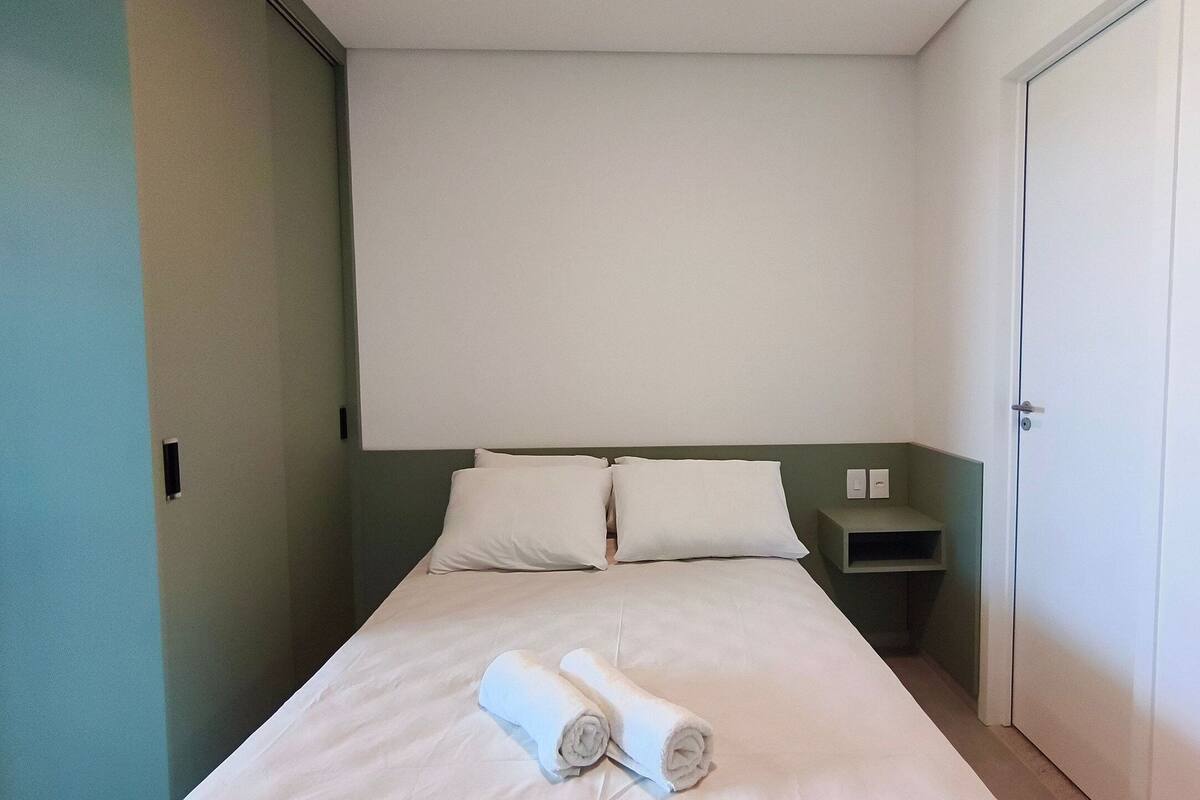 Un lit simple avec deux oreillers, une table de chevet, un interrupteur fixé au mur et une porte.
