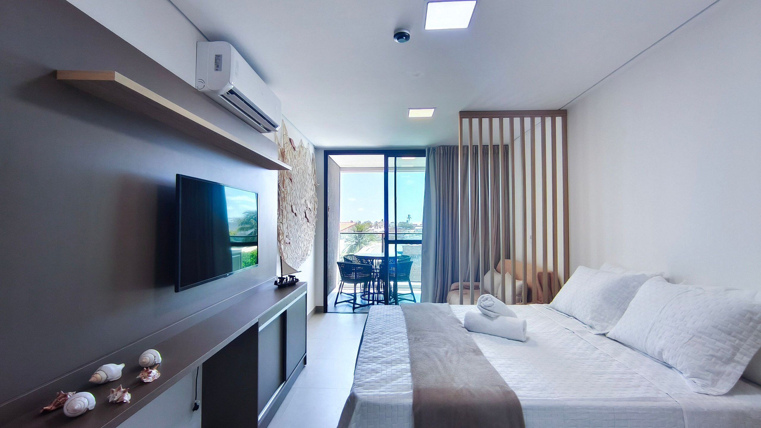 A modern hotel room with a flat-screen TV, a bed, a balcony with a table and chairs, and a view of the cityscape.