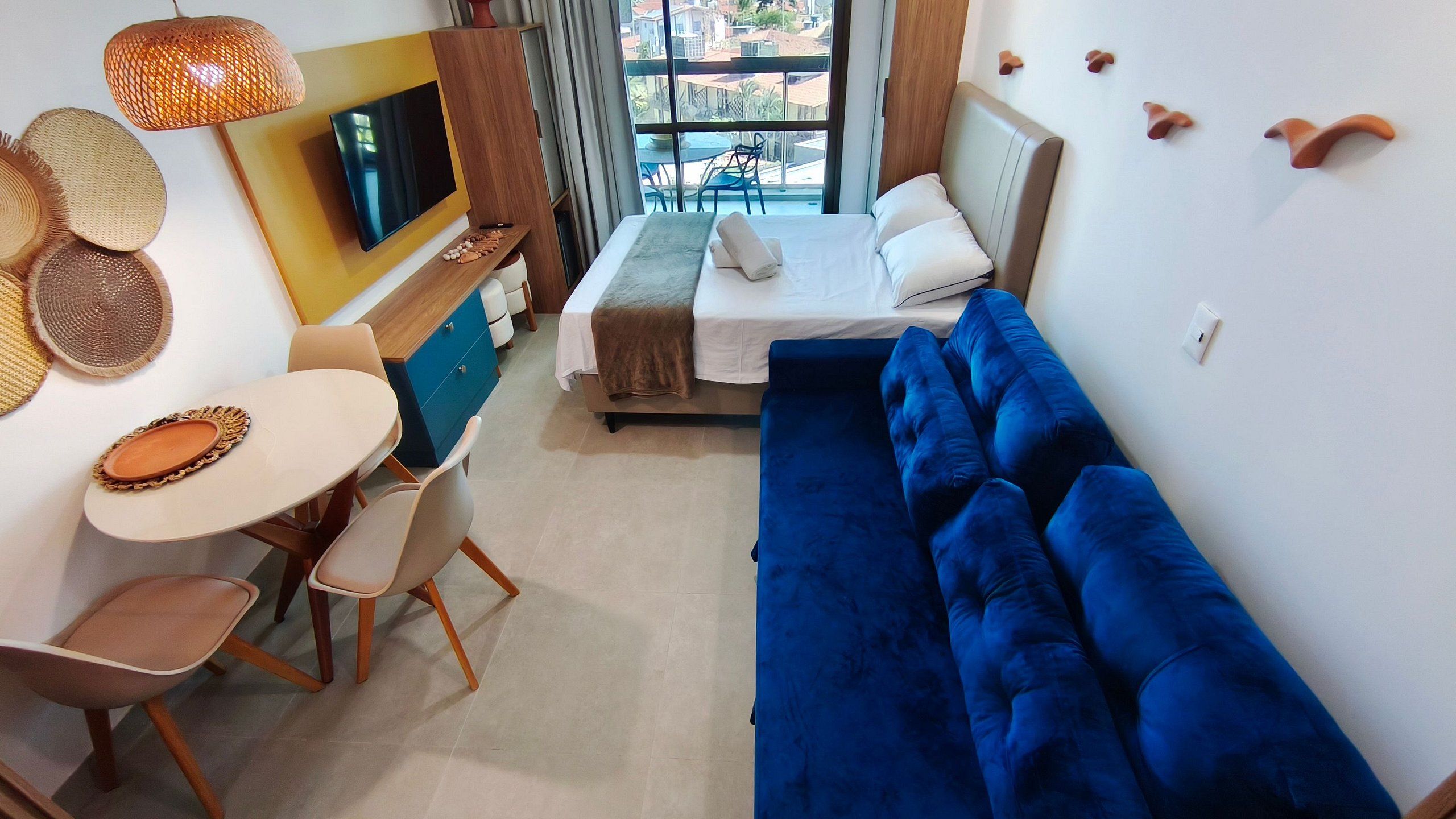 A modern hotel room with a bed, a blue sofa, a dining area with a table and chairs, a TV, and a view of the outside.