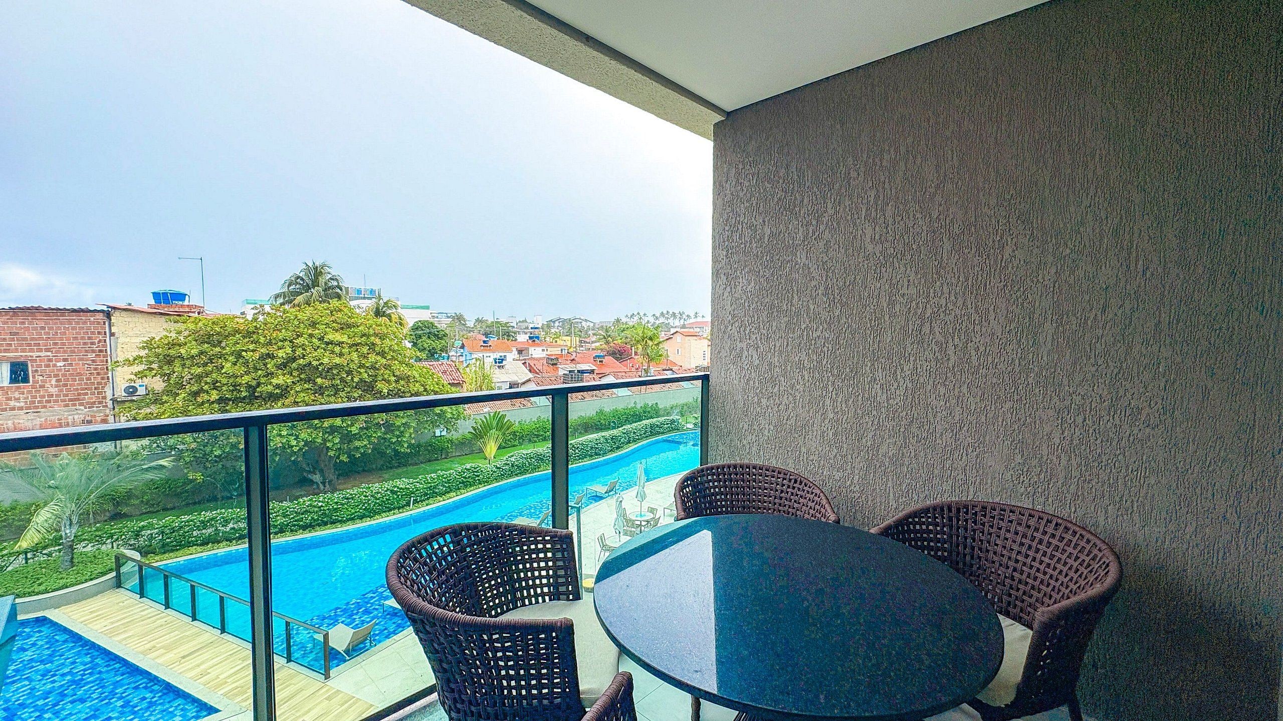Varanda com guarda-corpo de vidro, vista para uma piscina e edifícios.