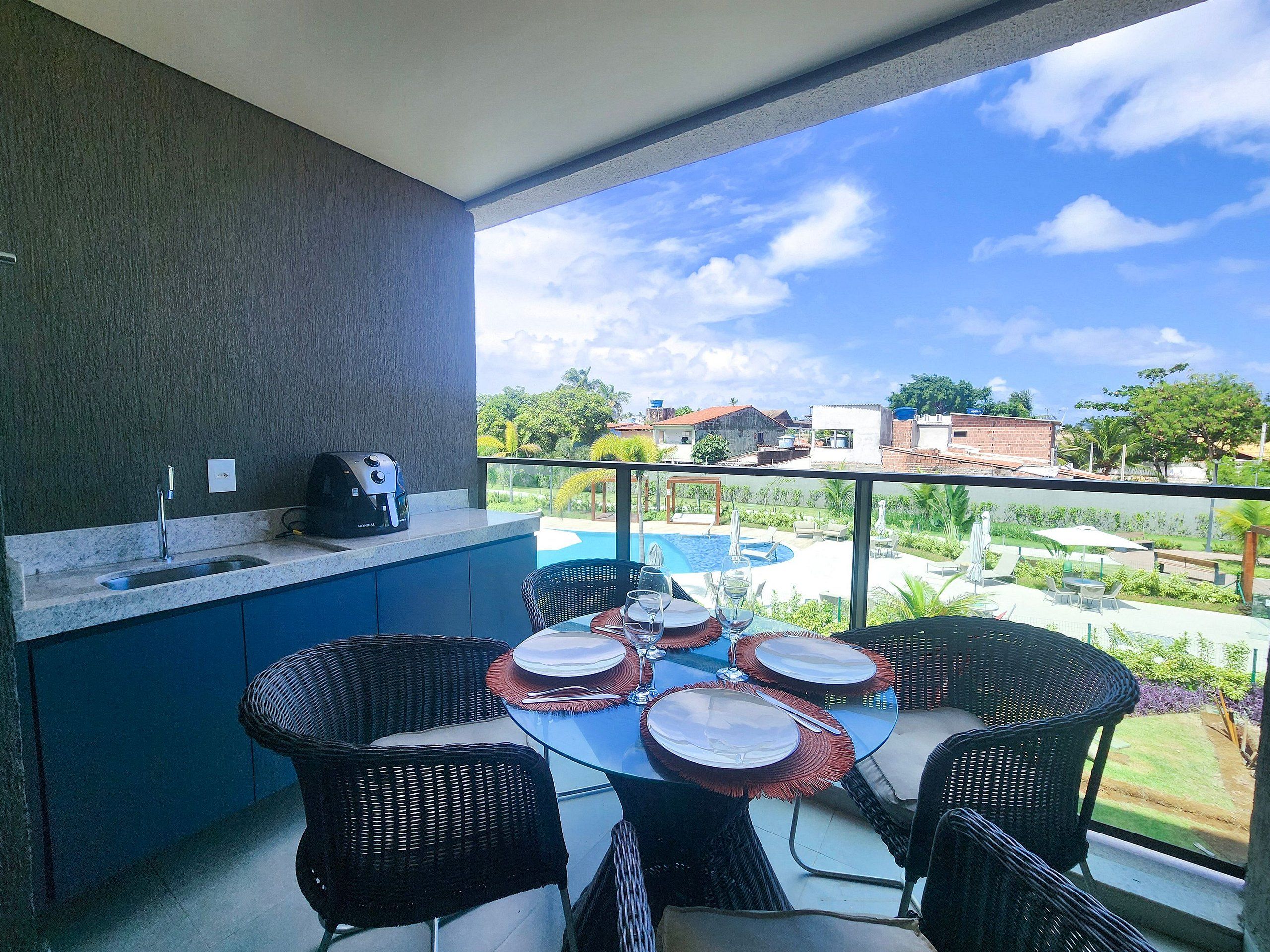 A balcony with a kitchen area, a dining table with chairs, and a view of a swimming pool and buildings.