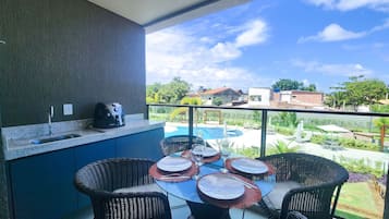 A balcony with a kitchen area, a dining table with chairs, and a view of a swimming pool and buildings.