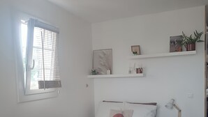 A bedroom with a bed, a bedside table with a lamp, a window with blinds, and shelves with decorative items.