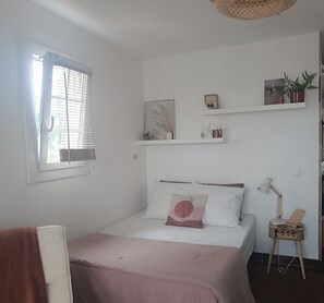 A bedroom with a bed, a bedside table with a lamp, a window with blinds, and shelves with decorative items. - Studio 🔆 just a few meters from the beach.  Ideal for couples or solo. (Agay)