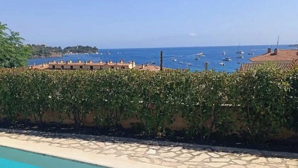 A swimming pool with clear blue water, a tiled deck, and a view of the sea with boats.