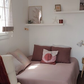 Une chambre à coucher comprenant un lit, des oreillers, une table de chevet, une lampe, une fenêtre avec des stores et une étagère fixée au mur, ornée d’objets décoratifs.