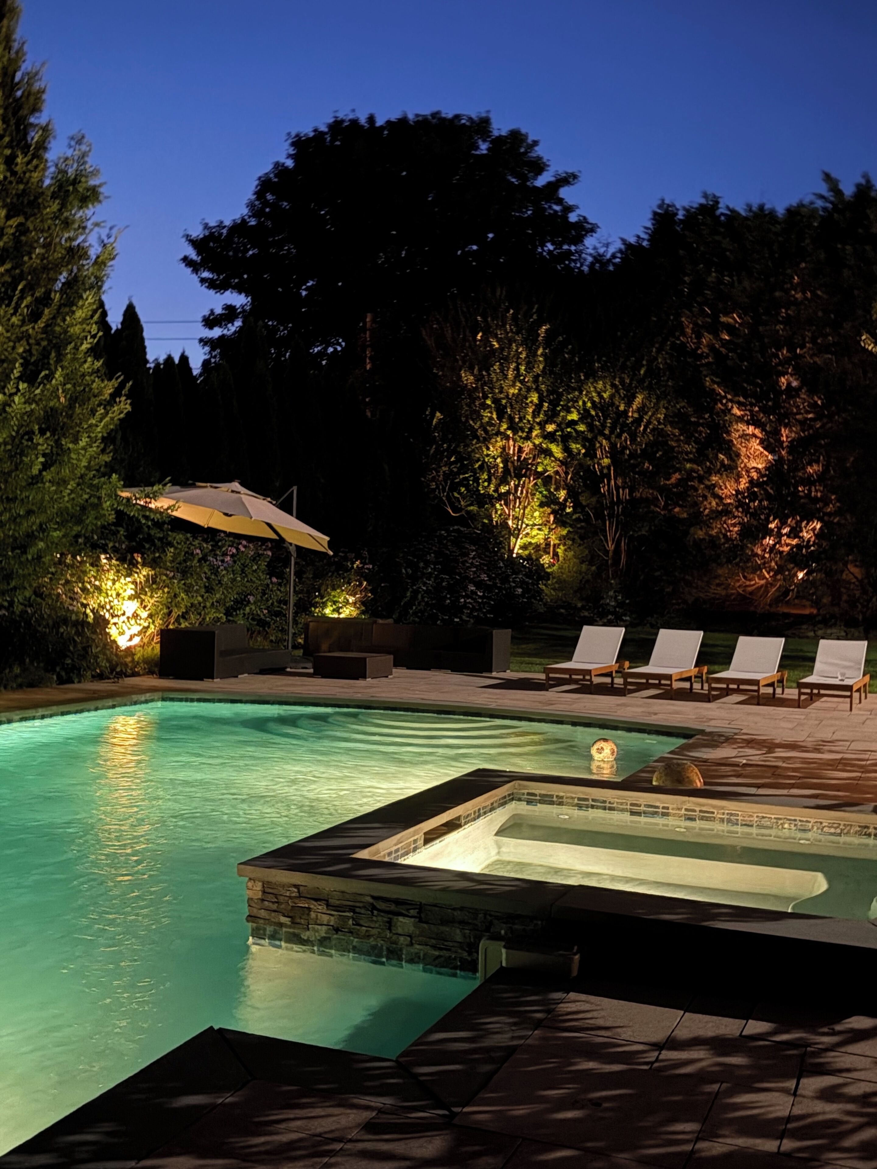 A swimming pool with lounge chairs and a hot tub, surrounded by trees and outdoor lighting.