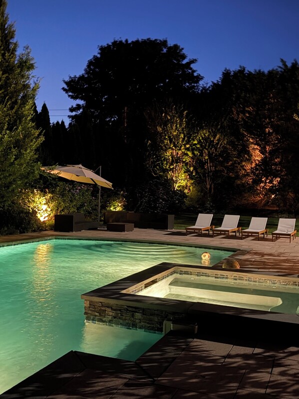 Piscina con sillas de descanso y jacuzzi, rodeada de árboles e iluminación exterior.