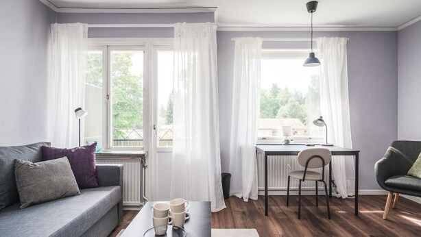 Un salon moderne avec un canapé, une table à manger et une chaise. On y trouve des fenêtres avec des rideaux, un radiateur et une lampe.