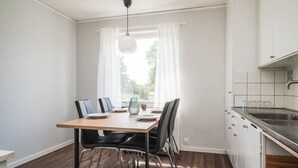 Un coin repas avec une table en bois, des chaises noires, un luminaire suspendu, une fenêtre avec des rideaux blancs et une cuisine dotée d’un évier en acier inoxydable.