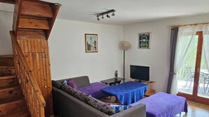 Una habitación con una escalera de madera, un sofá, una cama con cubrecama morado, un escritorio con ordenador y una ventana con cortinas.