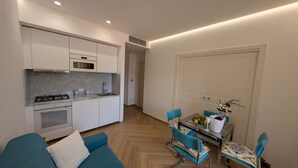 A compact kitchen with a stove, microwave, and sink, a dining area with a glass table and blue chairs, and a large white door. - Mondello D'Amare (Palermo)