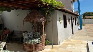A covered outdoor seating area with a wicker chair and a small table, a water tank on a building, and a clear blue sky.