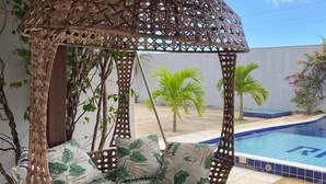 A wicker swing chair with tropical leaf cushions, a pool, and a pergola with climbing plants.