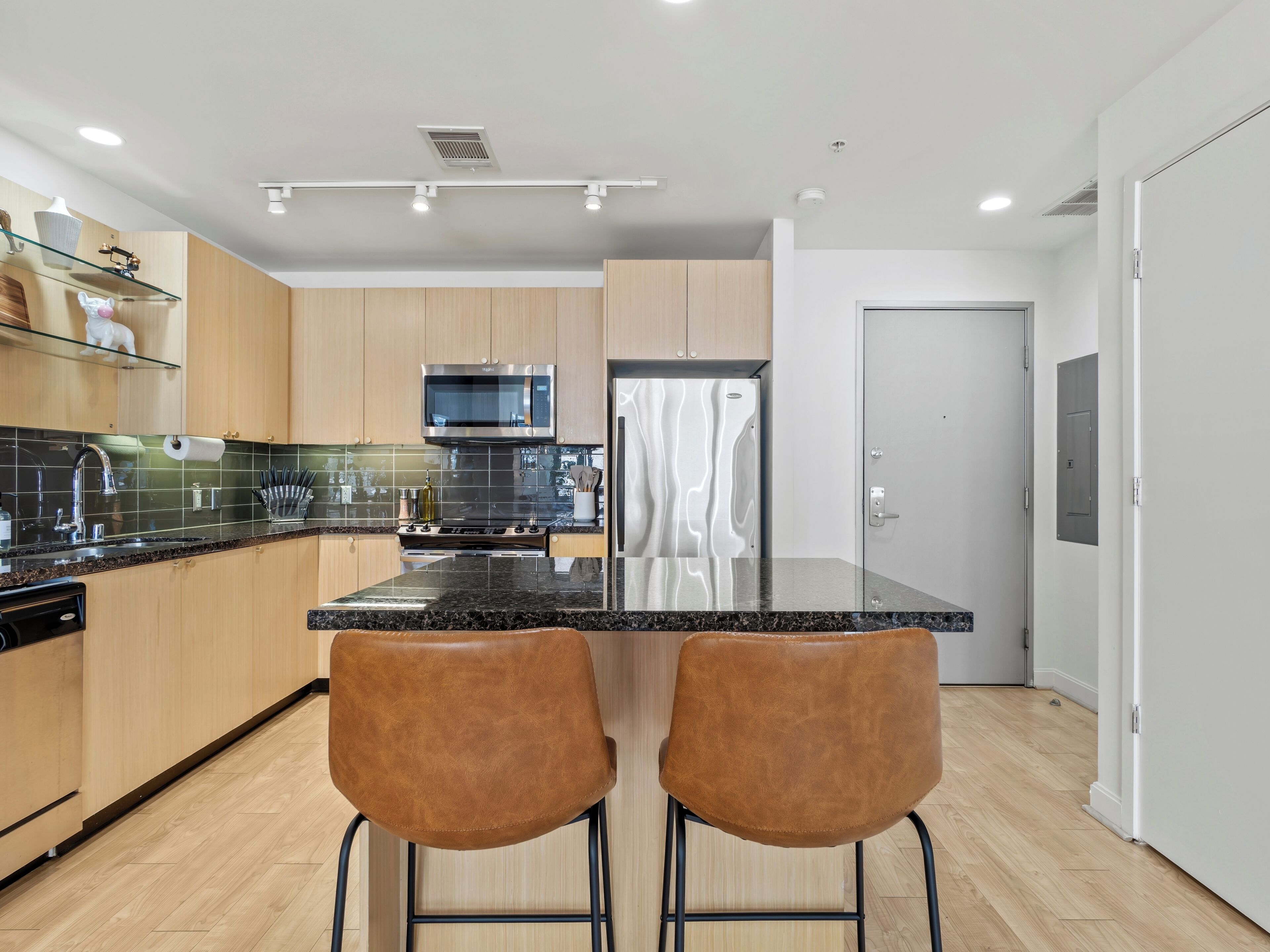 A modern kitchen with wooden cabinets, a black countertop, stainless steel appliances, and two brown bar stools.