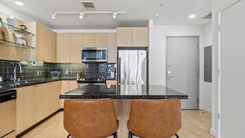 A modern kitchen with wooden cabinets, a black countertop, stainless steel appliances, and two brown bar stools.