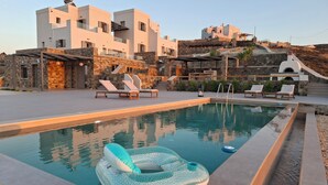 Uma área de piscina moderna com espreguiçadeiras e um flutuador, cercada por edifícios de pedra e uma encosta.
