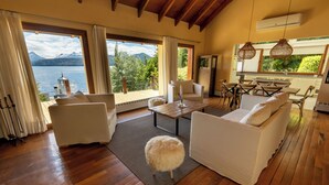 Un salón espacioso con suelo de madera, muebles blancos, un taburete mullido y un ventanal grande con vistas a un lago y montañas.