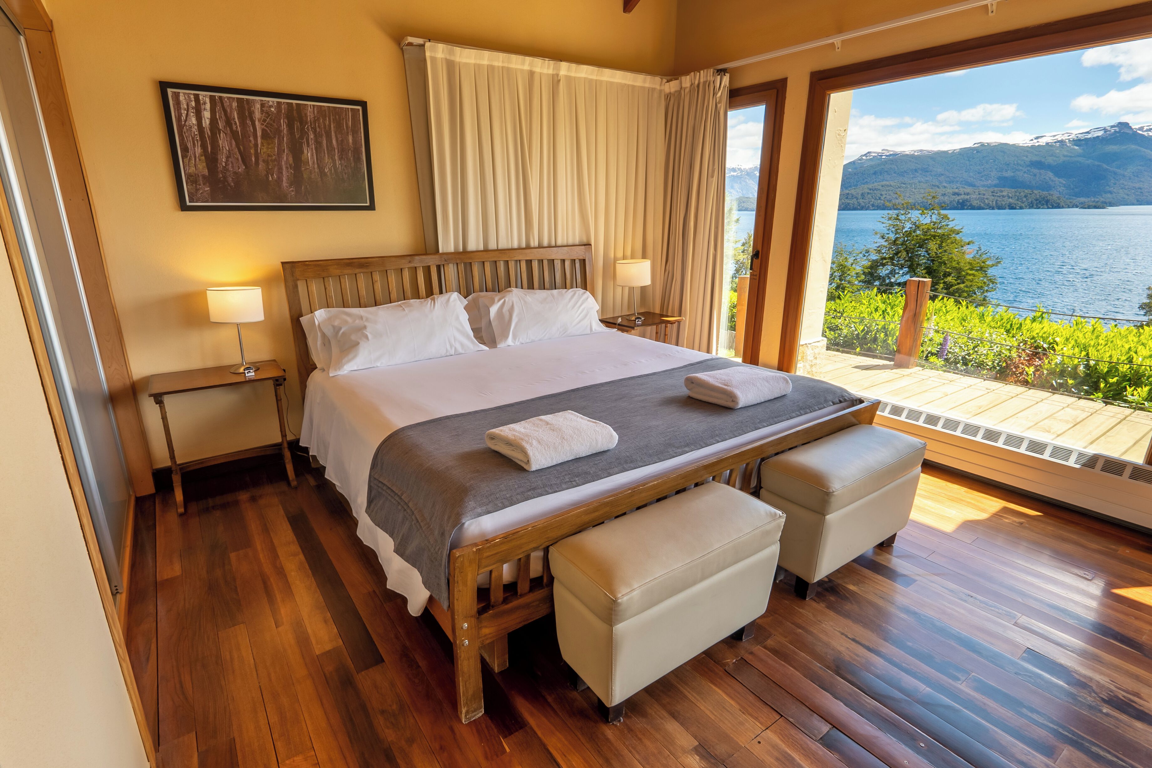A bedroom with a wooden bed, two bedside tables, a bench, and a view of a lake and mountains.