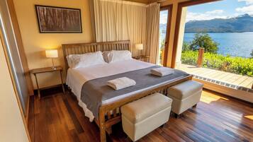 A bedroom with a wooden bed, two bedside tables, a bench, and a view of a lake and mountains.