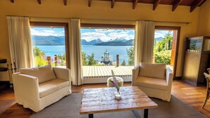 Una habitación con dos sofás blancos frente a una mesa de madera con un florero, ventanas amplias con cortinas y vistas a un lago y montañas.