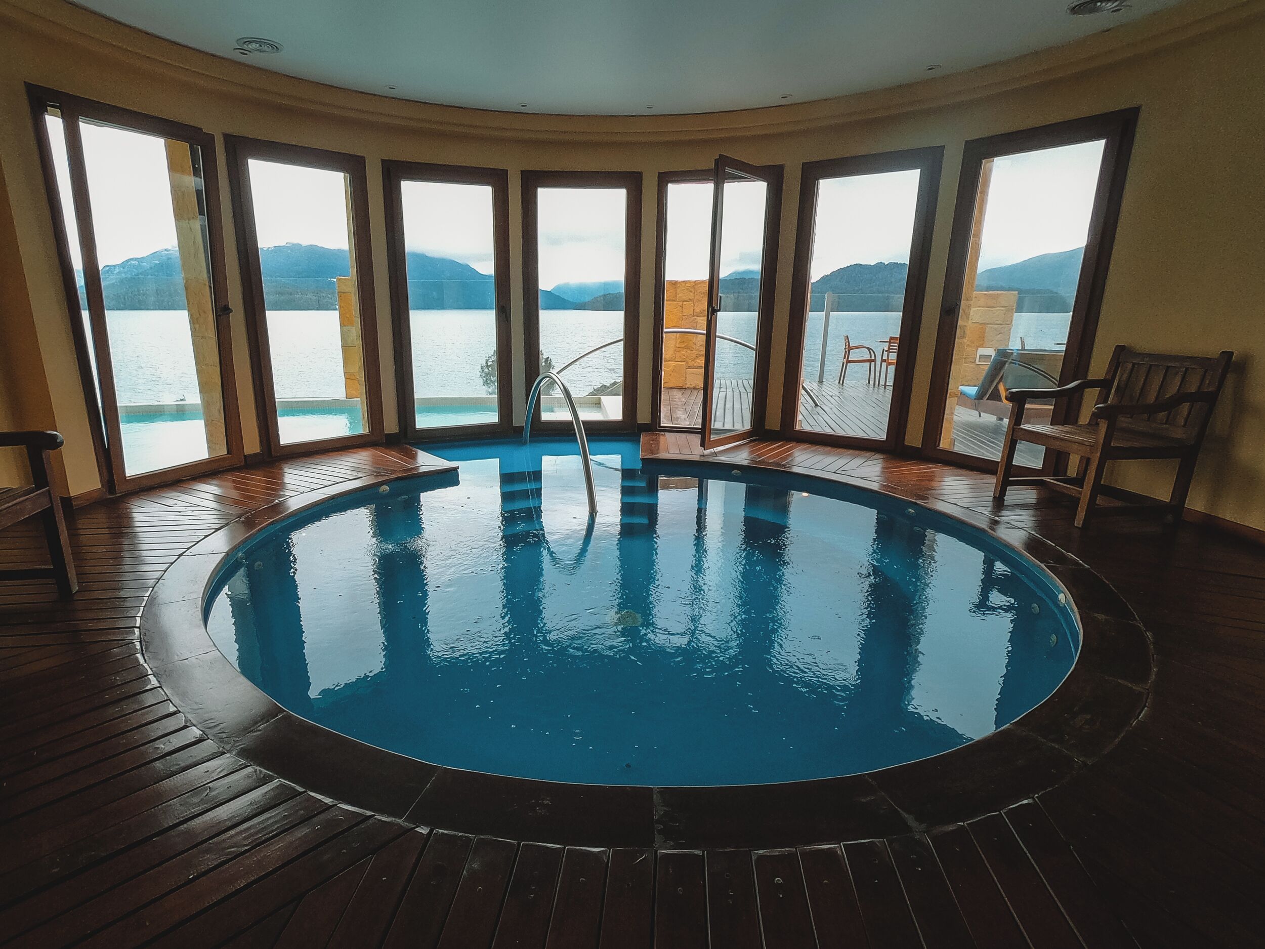 A circular indoor pool with a wooden deck surrounding it, large windows offering a view of a lake and mountains, and a few wooden chairs near the pool.