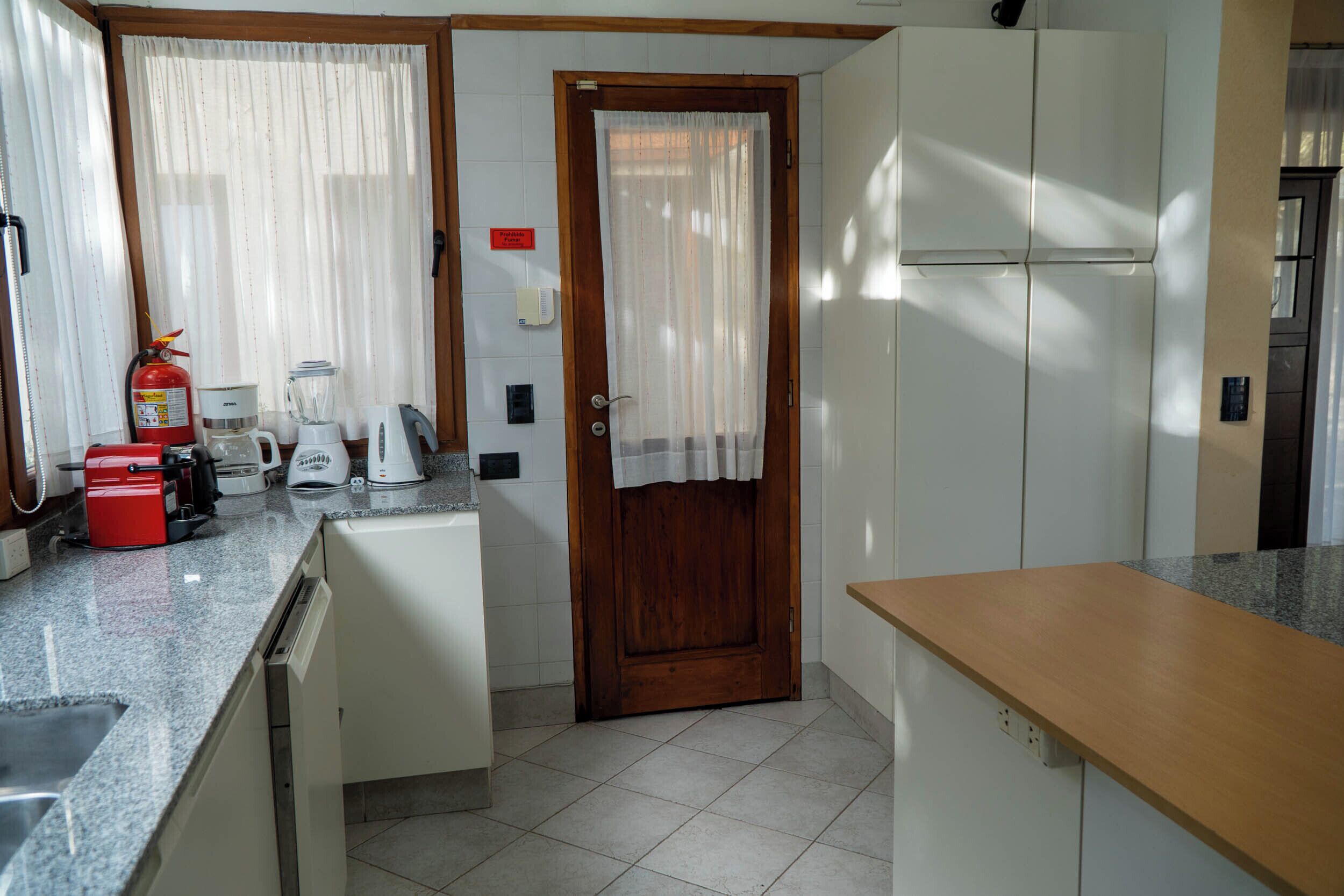 Una cocina compacta con puerta de madera, armarios blancos, una encimera con cafetera, licuadora y un tostador rojo.