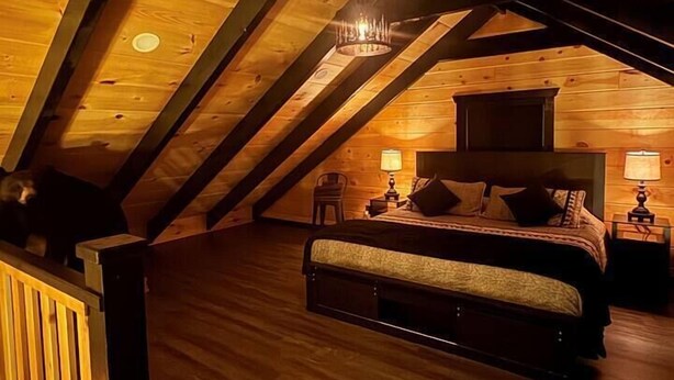 An attic bedroom with a wooden ceiling, a bed, two bedside tables with lamps, and a chandelier. - Cozy cabin with in the heart of Benezette, 1 mile from viewing center. (Benezette)