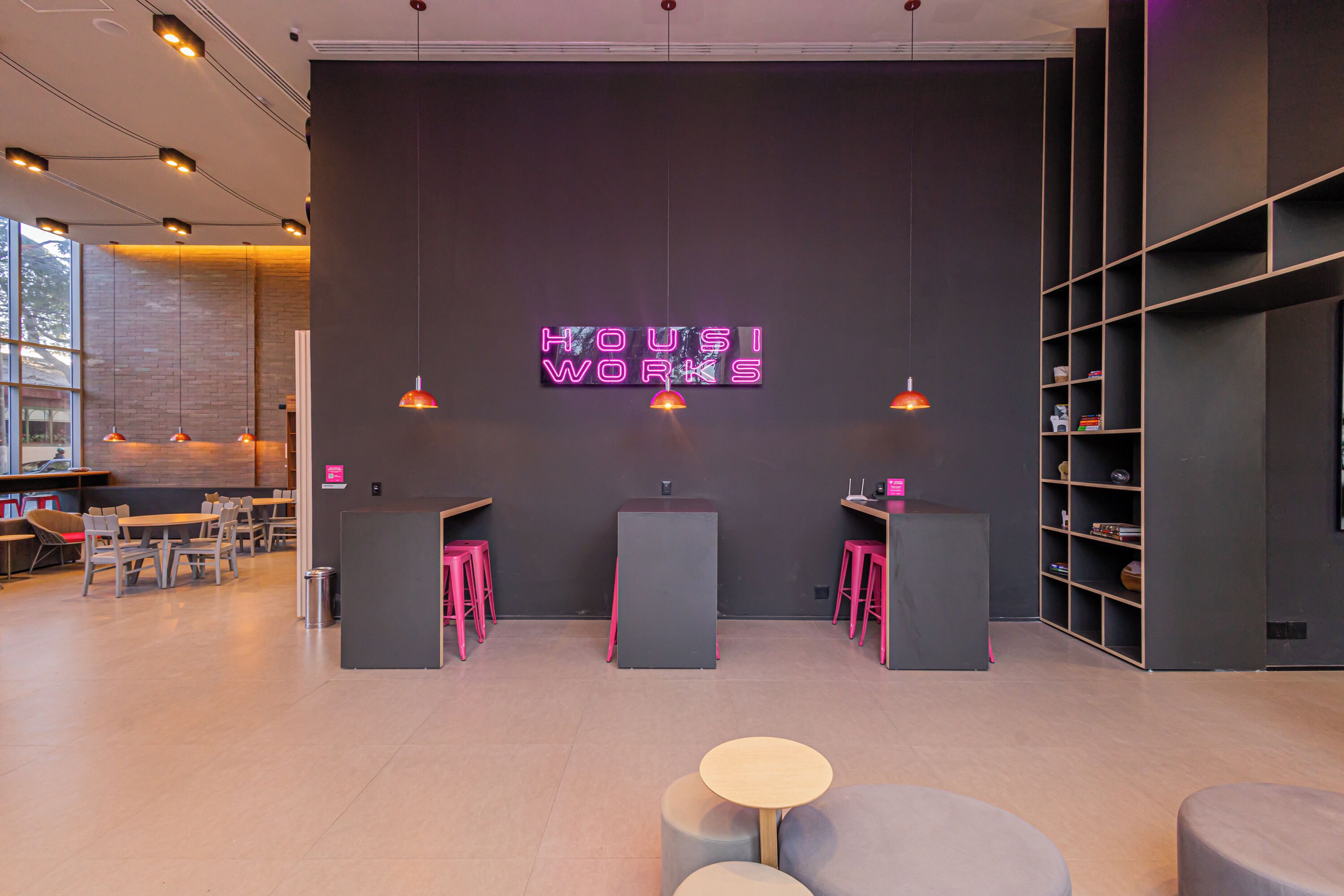 A modern interior with a neon sign reading "HOUSI WORKS," tables, chairs, and a bookshelf.