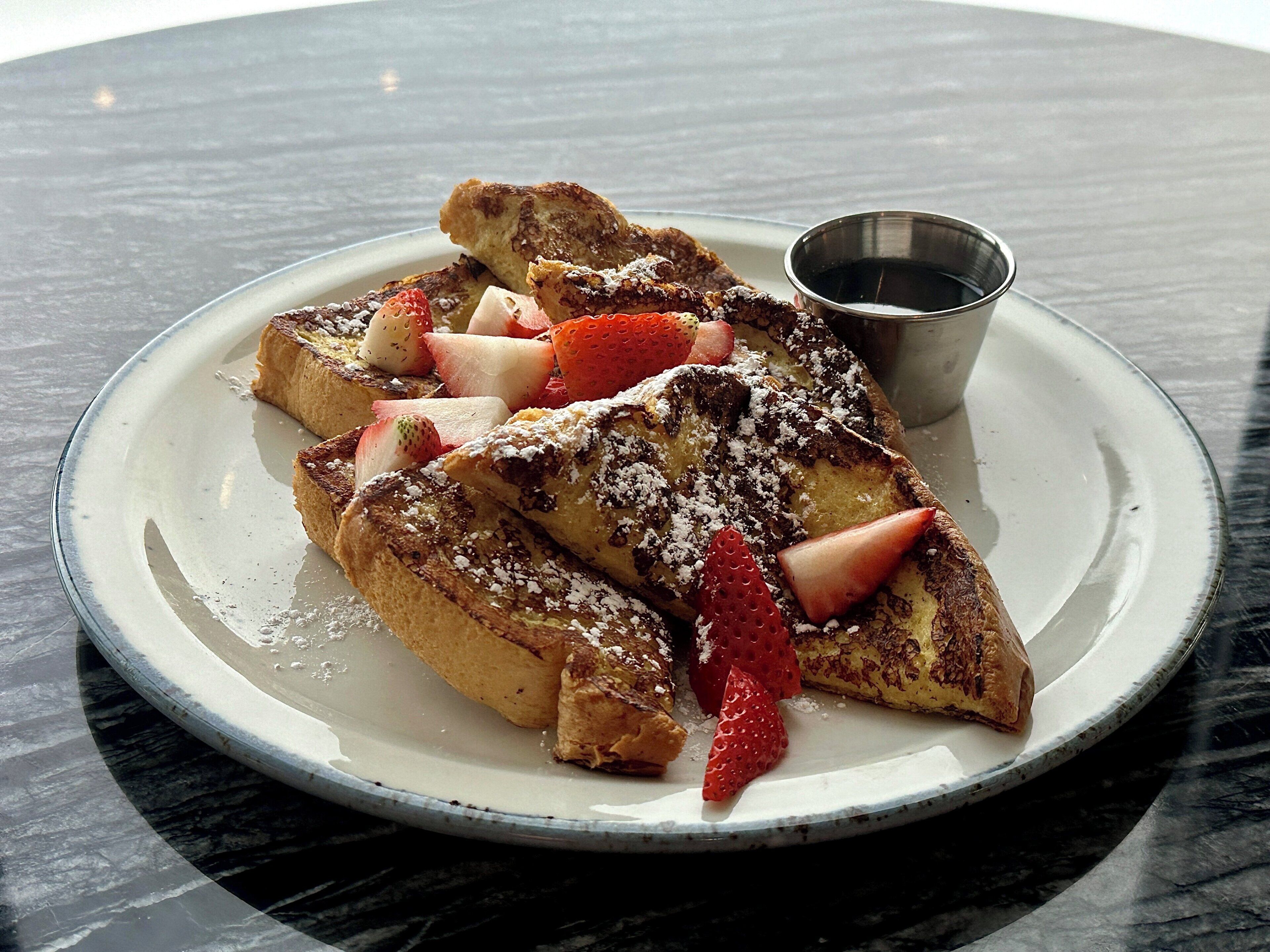 A plate of French toast with strawberries and powdered sugar, accompanied by a small metal cup, on a marble table.