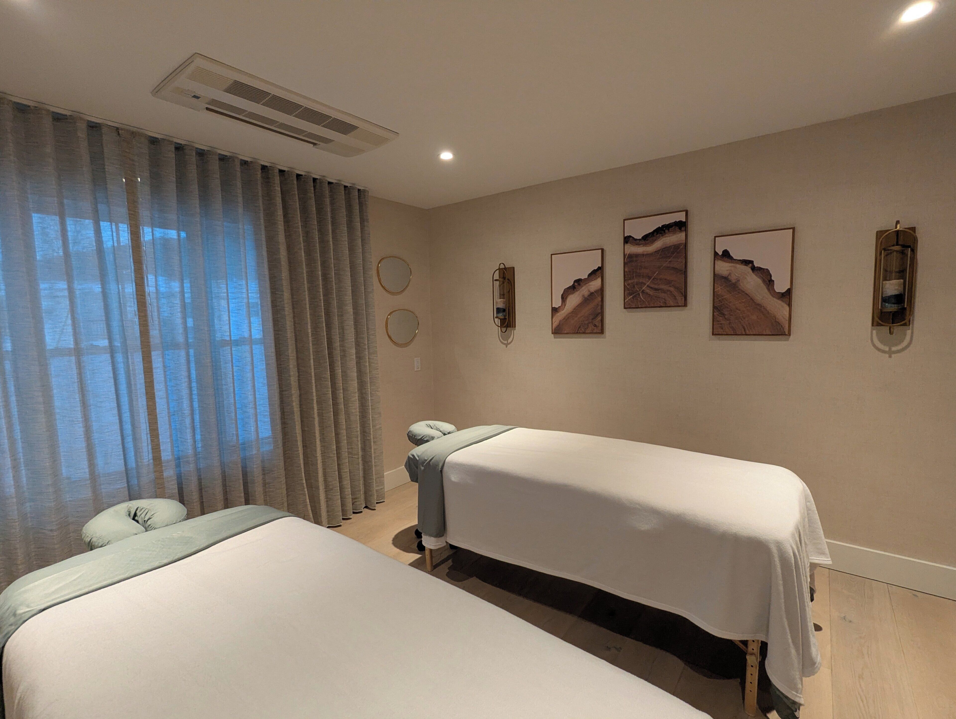 A hotel room with two beds, a large window with curtains, wall-mounted artwork, and a ceiling vent.