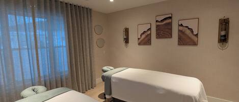 A hotel room with two beds, a large window with curtains, wall-mounted artwork, and a ceiling vent.