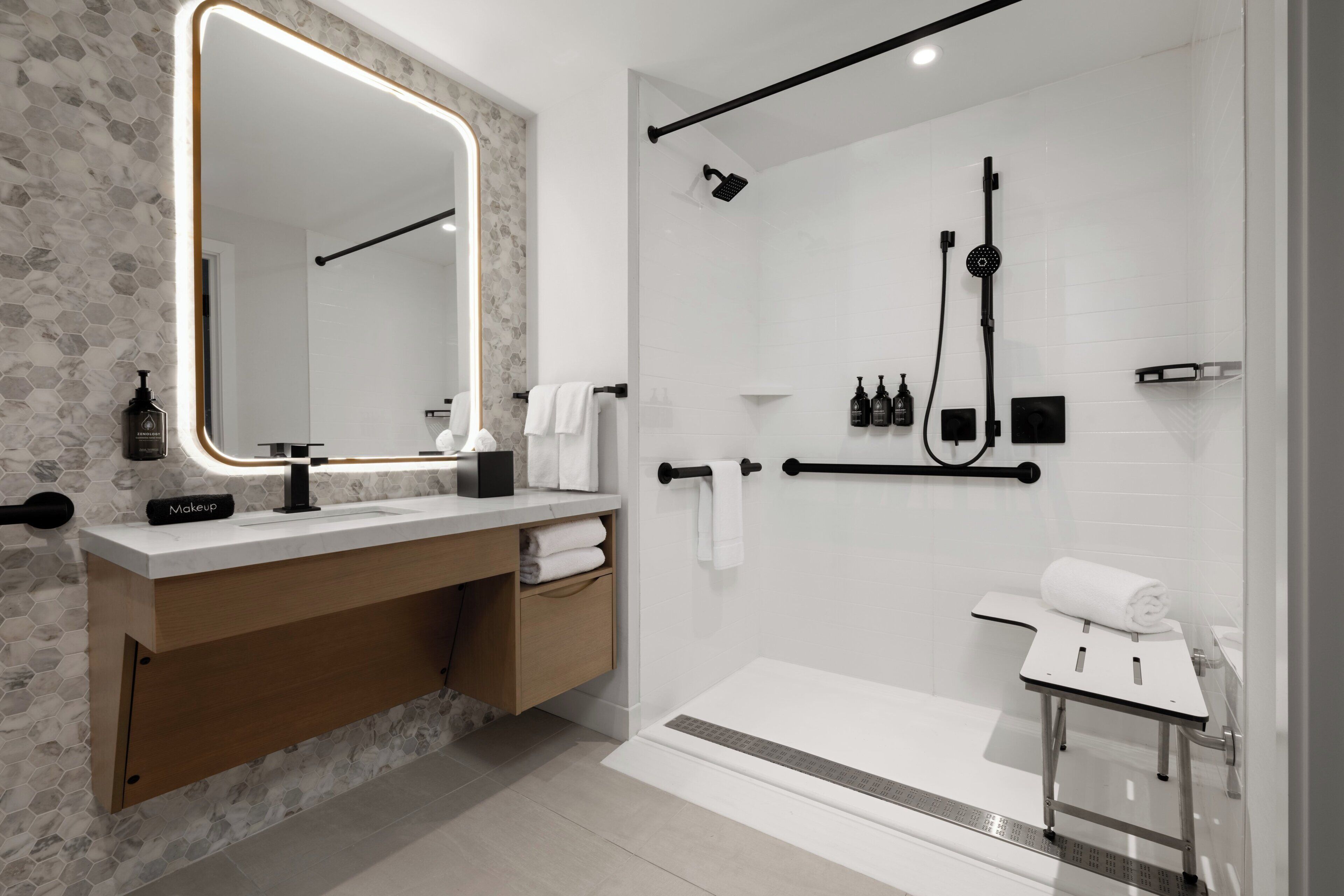 A modern bathroom with a large mirror, a vanity with a makeup station, a shower with a handheld showerhead, and a towel rack.