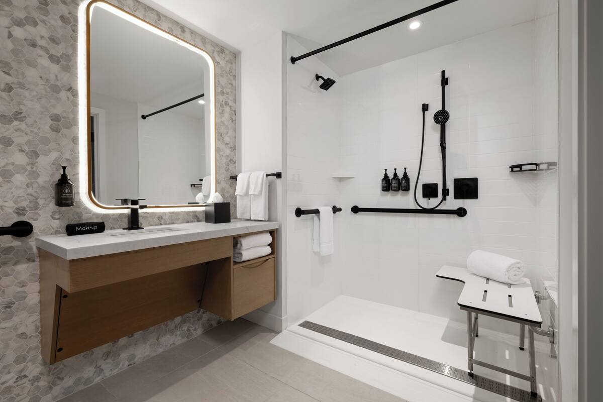 A modern bathroom with a large mirror, a vanity with a makeup station, a shower with a handheld showerhead, and a towel rack.
