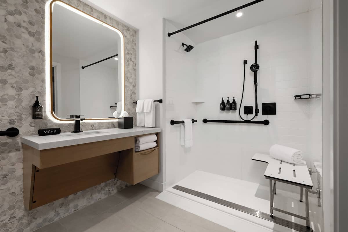A modern bathroom with a large mirror, a vanity with a makeup station, a shower with a handheld showerhead, and a towel rack.