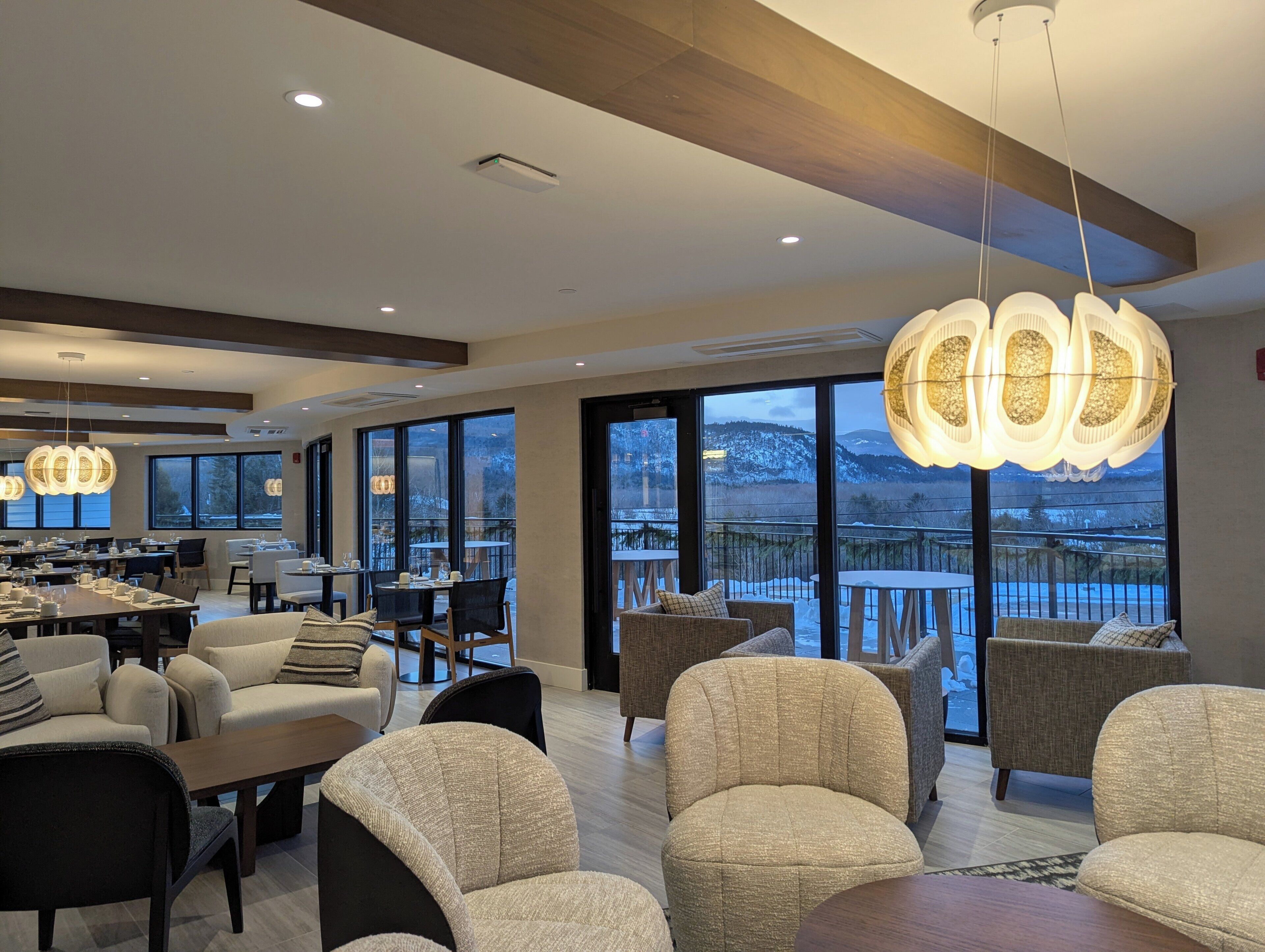 A modern hotel lobby with a dining area, comfortable seating, large windows with a view of a snowy landscape, and contemporary lighting fixtures.