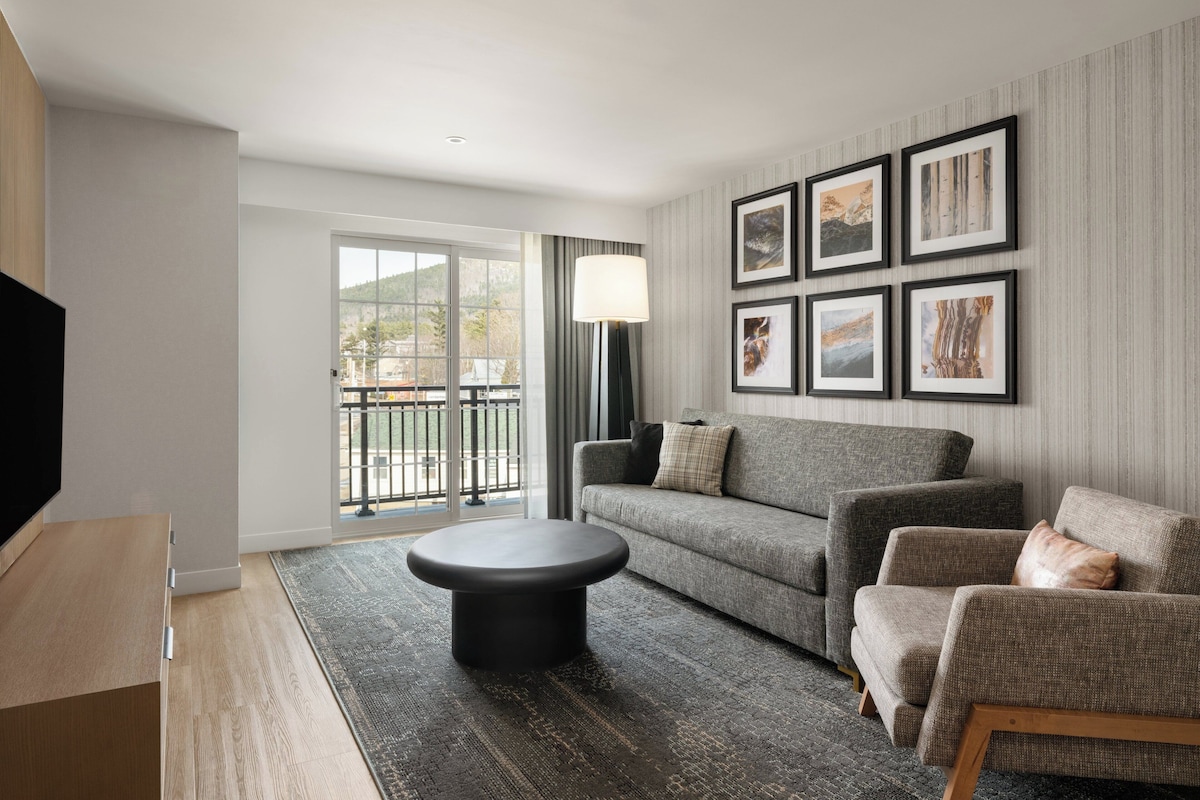 Una sala de estar moderna con sofá, sillón y reposapiés, un televisor de pantalla plana y un ventanal con vistas.