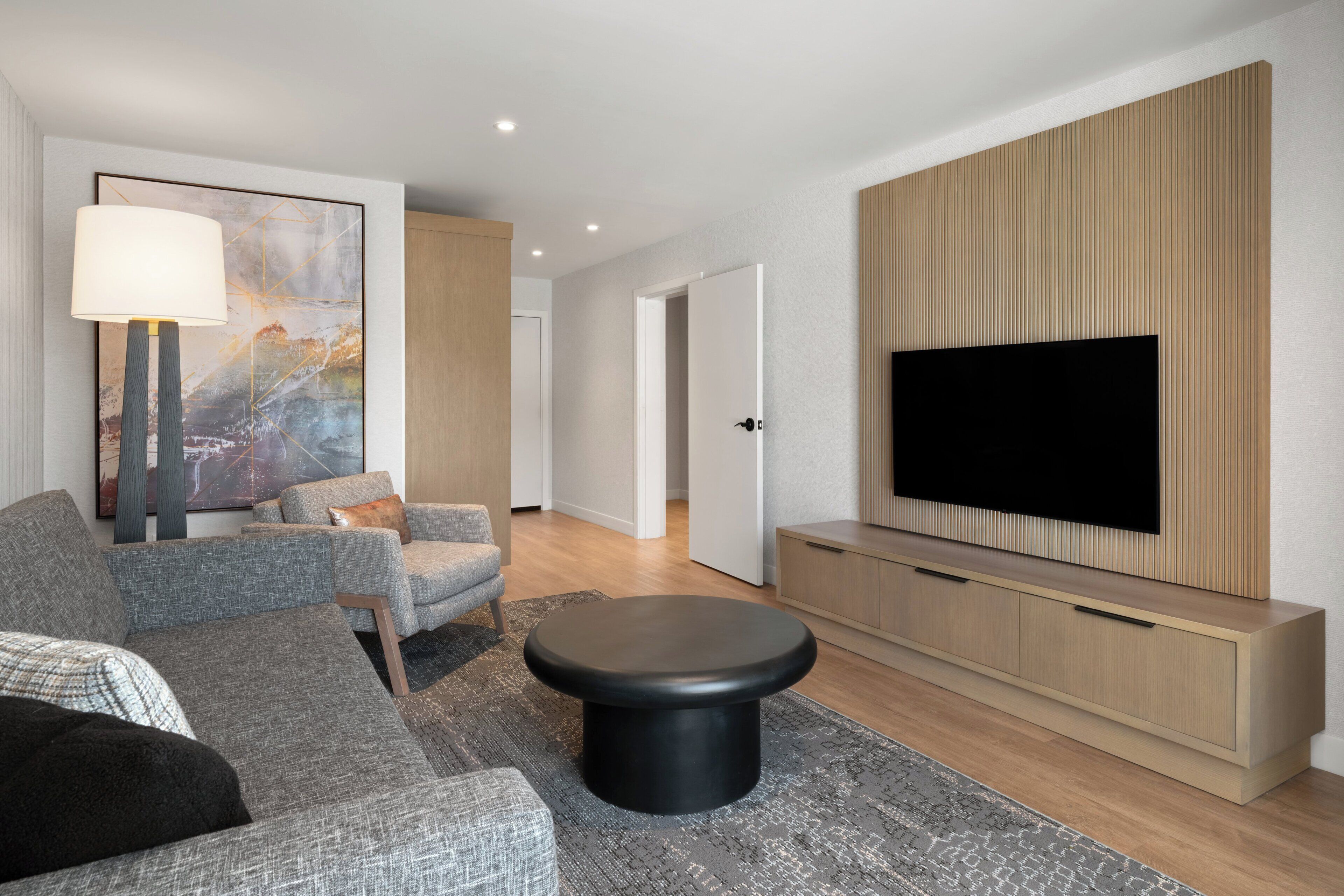A modern living room with a flat-screen TV, grey sofas, a black ottoman, and a large abstract painting.