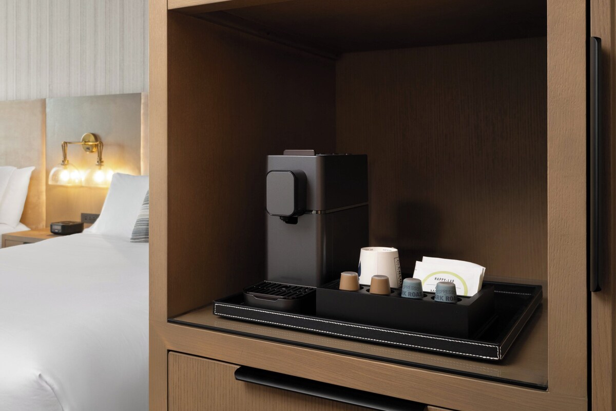 Una habitación de hotel con cama, mesita de noche, lámpara, cafetera y bandeja con tazas de café y una tarjeta.