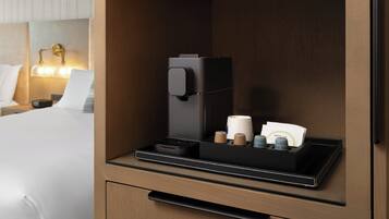 A hotel room with a bed, a nightstand, a lamp, a coffee machine, and a tray with coffee cups and a card.